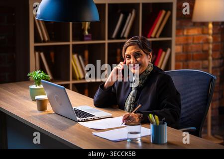 Senior Indian businesswoman in corporate attire taking notes while speaking on smartphone seated at workstation with laptop in cozy office space exper Stock Photo