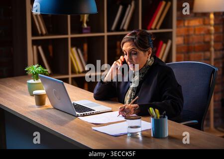 Senior Indian businesswoman in corporate attire taking notes while speaking on smartphone seated at workstation with laptop in cozy office space exper Stock Photo