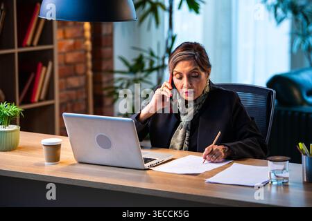 Senior Indian businesswoman in corporate attire taking notes while speaking on smartphone seated at workstation with laptop in cozy office space exper Stock Photo