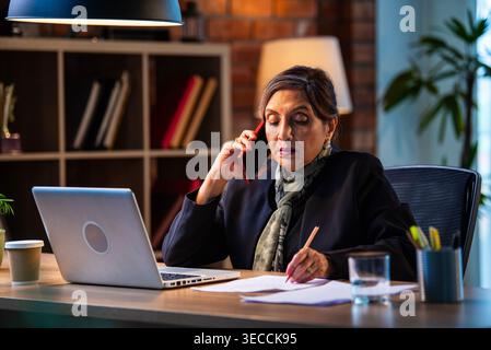 Senior Indian businesswoman in corporate attire taking notes while speaking on smartphone seated at workstation with laptop in cozy office space exper Stock Photo