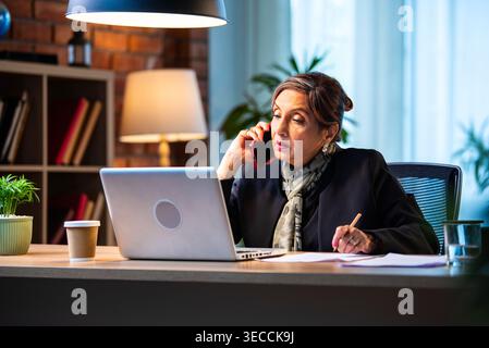 Senior Indian businesswoman in corporate attire taking notes while speaking on smartphone seated at workstation with laptop in cozy office space exper Stock Photo