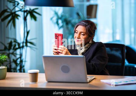 Senior Indian businesswoman in corporate attire chatting and typing on smartphone while sitting at workstation with laptop in modern cozy office space Stock Photo