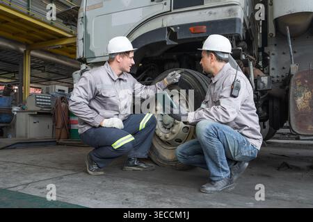 Caucasian and Middle Eastern mid-adult male technicians inspect a heavy truck tire using a digital tablet in an Asian shipyard, representing quality c Stock Photo