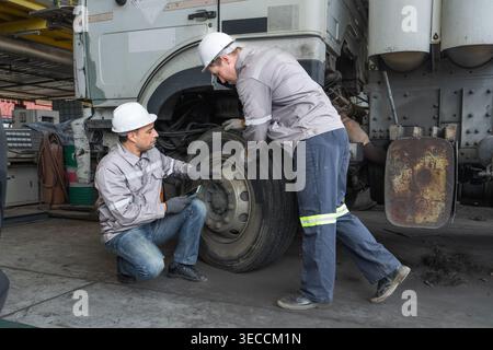 Caucasian and Middle Eastern mid-adult male technicians inspect a heavy truck tire using a digital tablet in an Asian shipyard, representing quality c Stock Photo