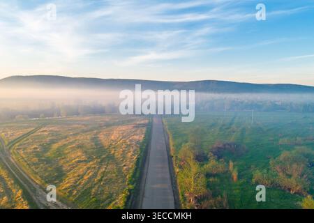 Aerial view captures long country road through sunlit fields, morning mist filling the valley, distant hills under blue sky Stock Photo