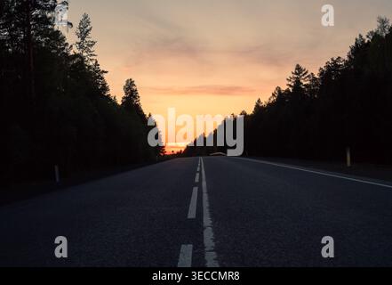 Empty highway road leading through a dense forest under a vibrant orange sunset sky Stock Photo