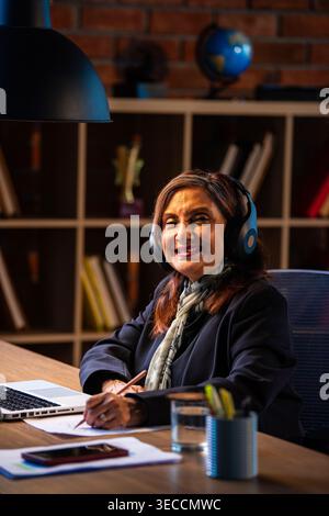 Senior Indian businesswoman in corporate attire taking notes while working on laptop with headphones at workstation in cozy office space experienced f Stock Photo