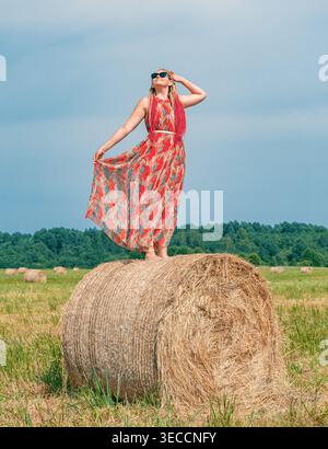 Woman standing on a large hay bale in a summer field, wearing a flowing dress and sunglasses under a bright sky Stock Photo