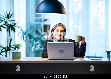 Senior Indian businesswoman in corporate attire showing coffee mug to camera with smile while sitting at work desk with laptop in cozy office space ex Stock Photo
