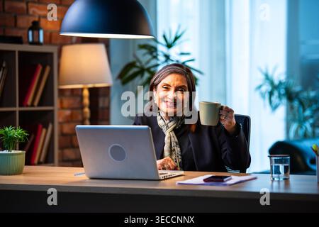Senior Indian businesswoman in corporate attire showing coffee mug to camera with smile while sitting at work desk with laptop in cozy office space ex Stock Photo