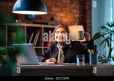 Senior Indian businesswoman in corporate attire showing coffee mug to camera with smile while sitting at work desk with laptop in cozy office space ex Stock Photo