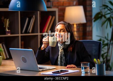 Senior Indian businesswoman in corporate attire showing coffee mug to camera with smile while sitting at work desk with laptop in cozy office space ex Stock Photo