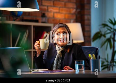 Senior Indian businesswoman in corporate attire showing coffee mug to camera with smile while sitting at work desk with laptop in cozy office space ex Stock Photo