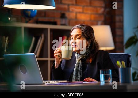 Senior Indian businesswoman in corporate attire showing coffee mug to camera with smile while sitting at work desk with laptop in cozy office space ex Stock Photo