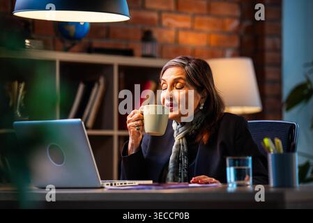 Senior Indian businesswoman in corporate attire showing coffee mug to camera with smile while sitting at work desk with laptop in cozy office space ex Stock Photo