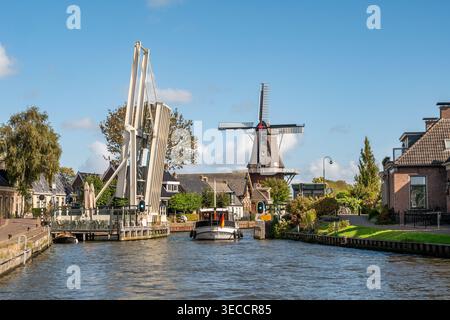 Motorboat passing open drawbridge over Dokkumer Ee canal and windmill De Zwaluw in village of Burdaard, Friesland, Netherlands Stock Photo