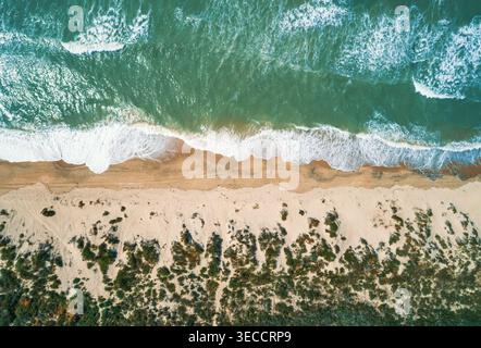 Aerial view shows vibrant green ocean water with white foam waves crashing onto a tan sandy beach. Stock Photo