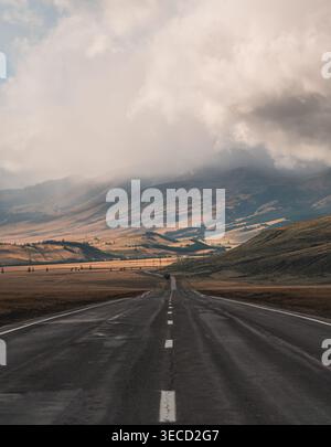 Straight highway through Altai plains extends toward distant mountains beneath a dark cloudy sky Stock Photo