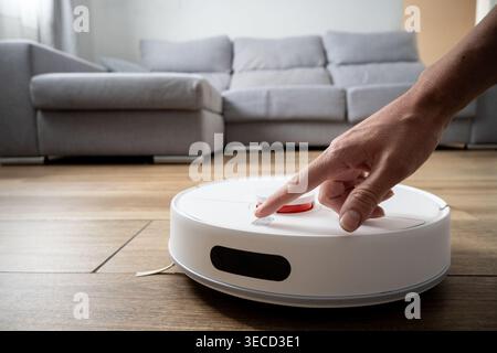 Close-up of a hand pressing the control button on a smart robot vacuum cleaner on a wooden floor. Concept of home automation, smart technology, user i Stock Photo