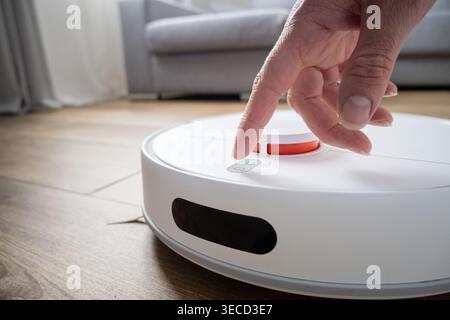 Close-up of a hand pressing the control button on a smart robot vacuum cleaner on a wooden floor. Concept of home automation, smart technology, user i Stock Photo