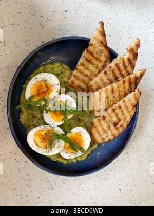 Smashed avocado topped with soft-boiled eggs, accompanied by grilled toast. Stock Photo