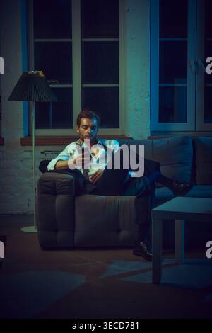 Man sitting on couch checking phone with serious expression in evening interior. Stock Photo