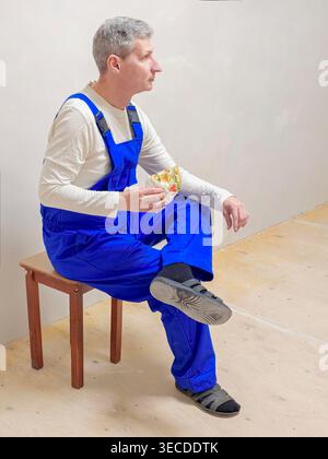 A break from work, a tea time. A worker in blue overalls is sitting on a stool, eating pizza. He looks thoughtful and relaxed, with a simple white background highlighting the scene. This would be ideal for images of workers taking a break. Stock Photo