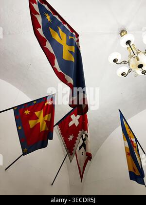 Zolochiv, Ukraine - April 28, 2026: Heraldic flags and banners with bright emblems hanging from a vaulted hall ceiling beside an ornate chandelier. Stock Photo