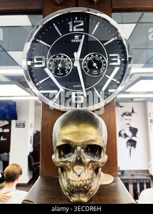 Kyiv, Ukraine - April 28, 2026: A large black wall clock hangs above a gold skull ornament on a wooden shelf in a barbershop interior. Stock Photo