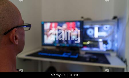 Man wearing glasses works at desk with dual monitors, one showing a speaker, the other a computer interior; home office setup, focused technology and Stock Photo