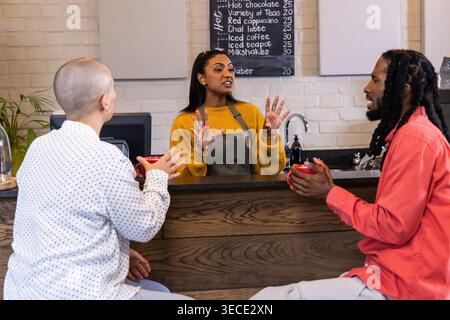 Female barista wearing gray apron, gesturing at coffee shop counter, customers holding red mugs. Cafe, warmth, cozy, conversation, chalkboard, brick, Stock Photo