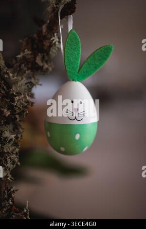 Easter bunny themed egg ornament hanging from a textured tree branch, featuring green felt ears, a hand-drawn face, and polka dots, symbolizing spring Stock Photo