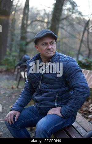 Middle-aged man sitting on a park bench in autumn, wearing a flat cap and blue quilted jacket, gazing seriously into the distance with a focused and s Stock Photo