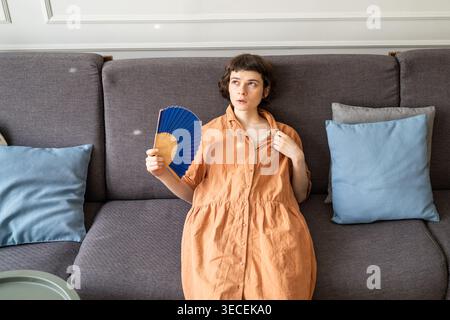Woman on couch with sick expression waving hand fan overwhelmed by summer heat in apartment. Stock Photo