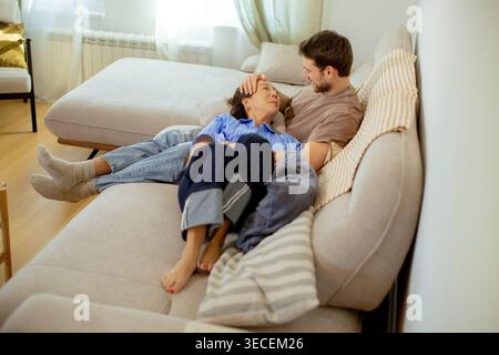 A Korean woman rests her head on the lap of a Caucasian man while they sit on a couch. They are in a living room with large windows. The atmosphere is Stock Photo