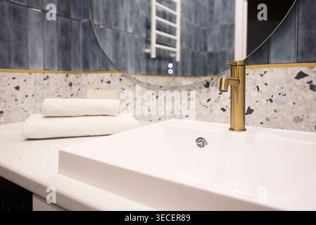 Contemporary bathroom features a white ceramic sink with a gold faucet and stacked white towels on a marble countertop, blue tiles in the background Stock Photo