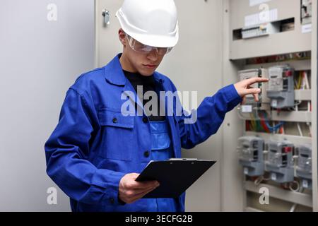 Electrician with clipboard checking electrical meters in fuse box indoors Stock Photo