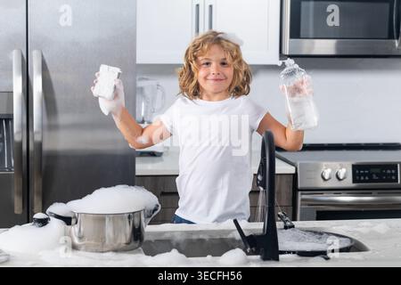 Boy washing dishes with bubbles. Child playing with foam in kitchen. C Funny boy enjoying house chores. Playful child washing kitchenware. Kid in soap Stock Photo