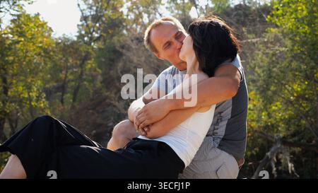 Happy couple hugs during sitting on wooden log on sandy beach. Romantic man kissing his beloved near coastline. Carefree lovers spending time together Stock Photo