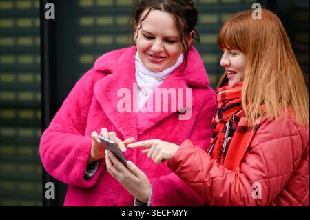 Two happy sisters using smartphone together on city street. Young women in bright pink, red coats browsing mobile app or choosing clothes online. Conc Stock Photo