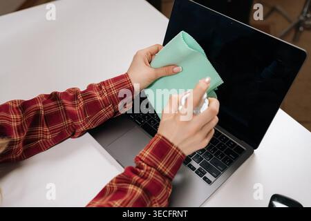 High angle view of female hands spraying cleaning solution on green cloth to wipe computer screen. Concept of hygiene, device care, and tidy workspace Stock Photo