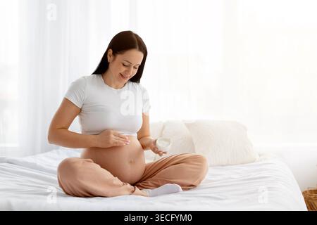 Young pregnant woman sitting on bed applying cream to belly Stock Photo