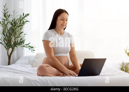 Smiling pregnant woman sitting on bed using laptop, browsing or working online Stock Photo