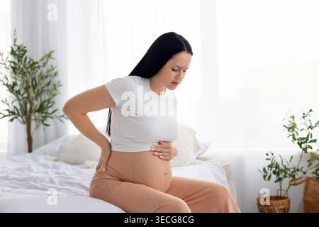 Pregnancy discomfort. Pregnant woman sitting on bed holding lower back in pain Stock Photo