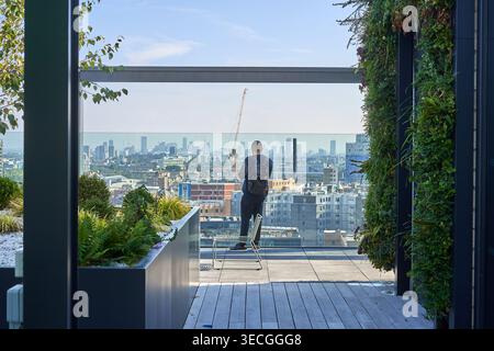 A man stands on a modern rooftop terrace overlooking the London cityscape, viewed from behind wearing dark clothing and a backpack. The contemporary Stock Photo
