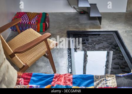 A mid-century modern wooden armchair with woven rope seating sits on a polished concrete floor next to a built-in rectangular water feature filled Stock Photo