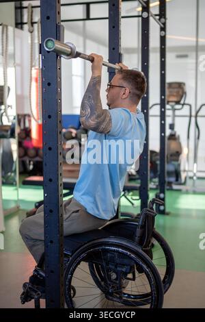 Man in wheelchair doing vertical pull ups workout using adaptive fitness equipment in sports center. Stock Photo