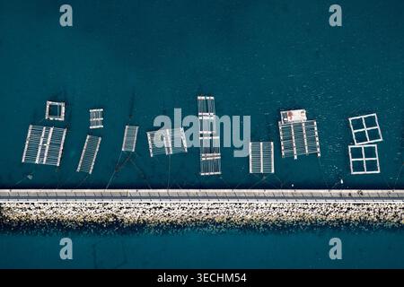 Aerial view of floating oyster farm structures and rectangular cages in the deep blue sea along a stone breakwater at Itoshima Peninsula Itoshima, Fuk Stock Photo