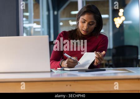 Indian woman sitting at wooden desk in office, wearing maroon top, writing in notebook near laptop Stock Photo