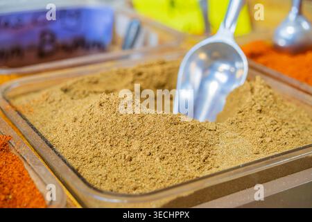 A silver scoop rests deeply embedded in a mound of fine, golden-brown spice powder inside a clear container at a market. Stock Photo